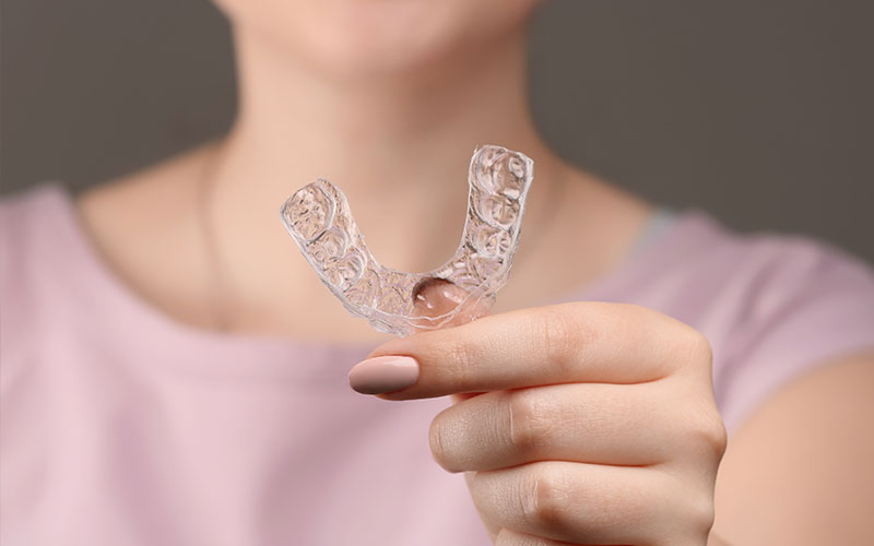The image shows a person s hand holding up a clear plastic dental impression, with the background blurred out.